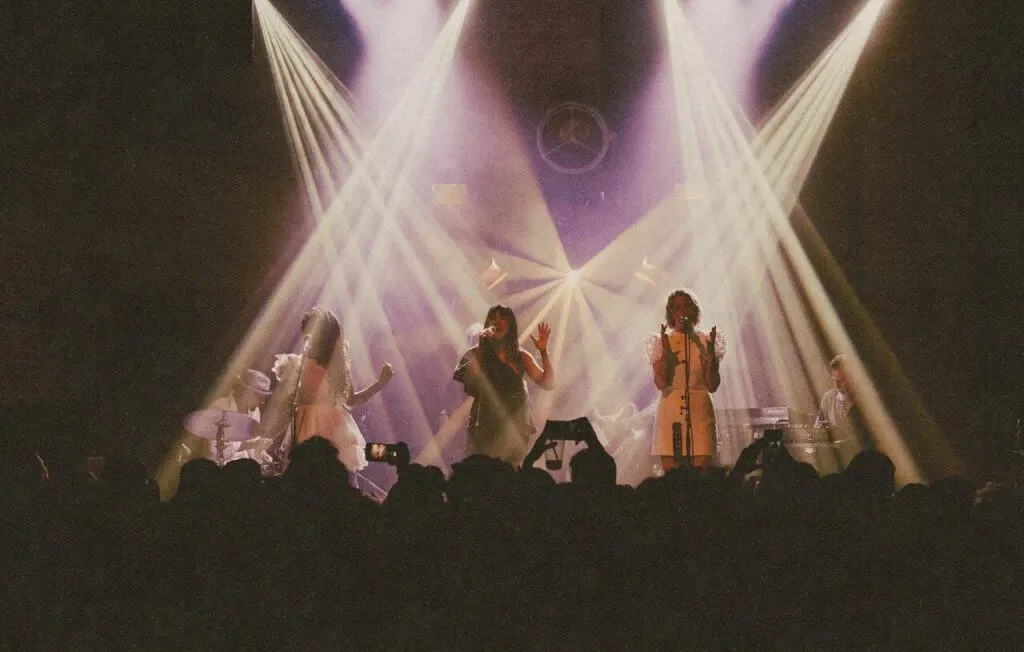 Live music performance with multiple performers on stage under dramatic purple and white stage lighting, audience members in foreground.