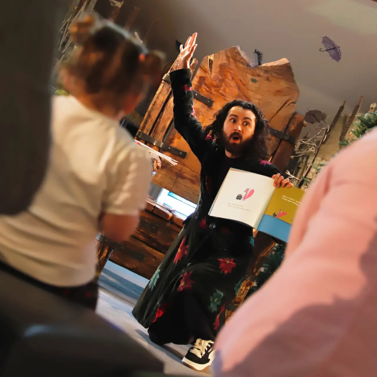 An enthusiastic presenter gestures expressively whilst holding artwork during an interactive storytelling session with children indoors.
