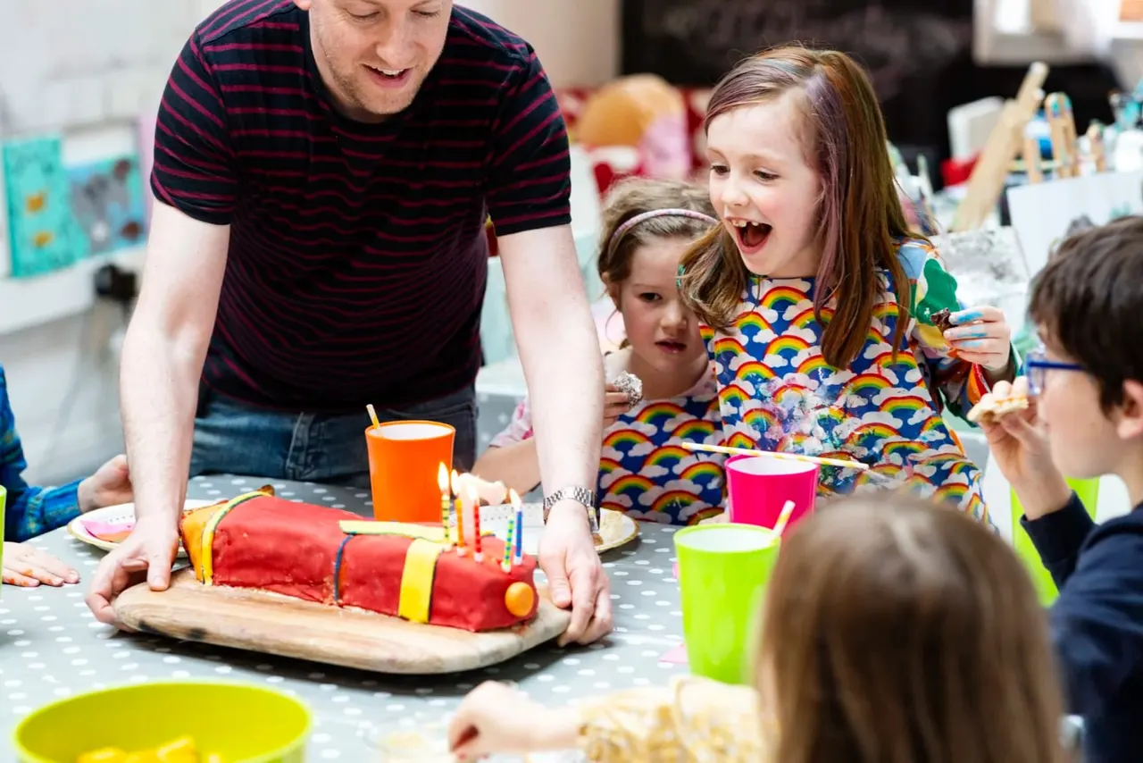 An adult and several young children gather around a table celebrating with a decorated cake and colourful cups at a birthday party.