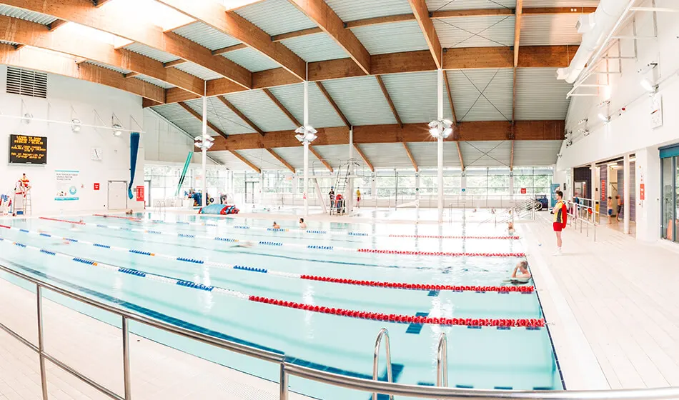 Indoor swimming pool with multiple lanes divided by blue and red lane ropes, featuring exposed wooden beams and metal roof structure.