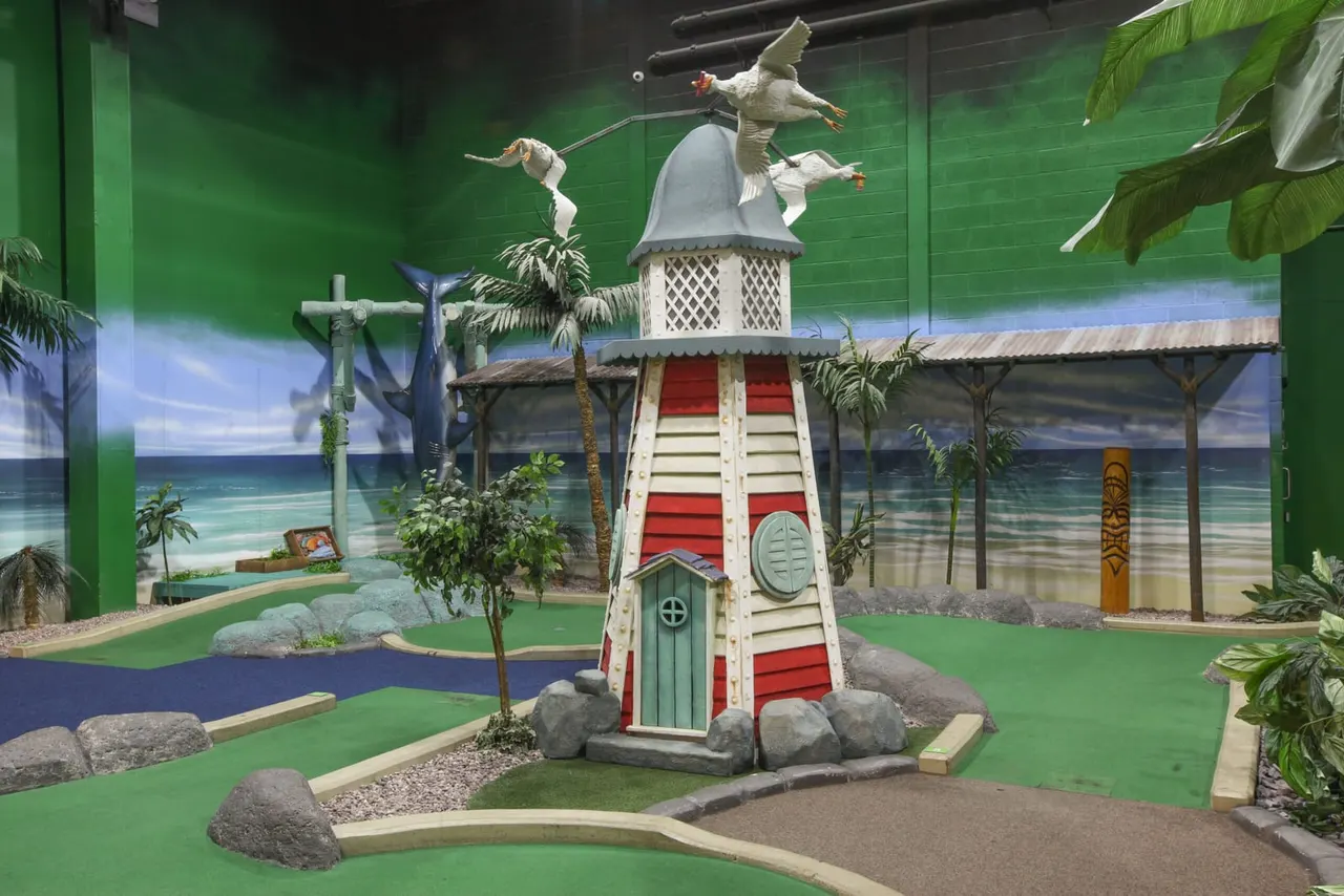 Indoor mini-golf course with a lighthouse-themed structure and tropical scenery backdrop.