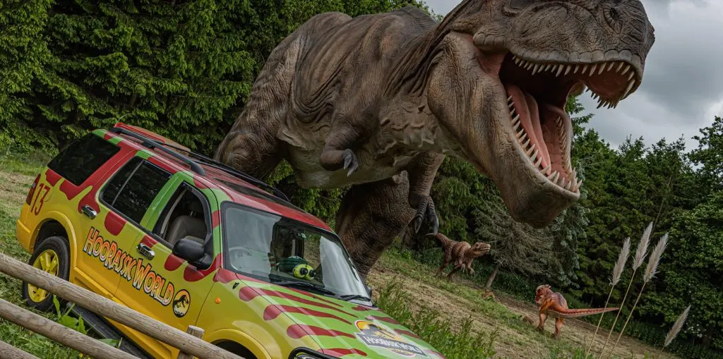Colourful tour vehicle beneath a large dinosaur model with open jaws in a leafy outdoor setting.