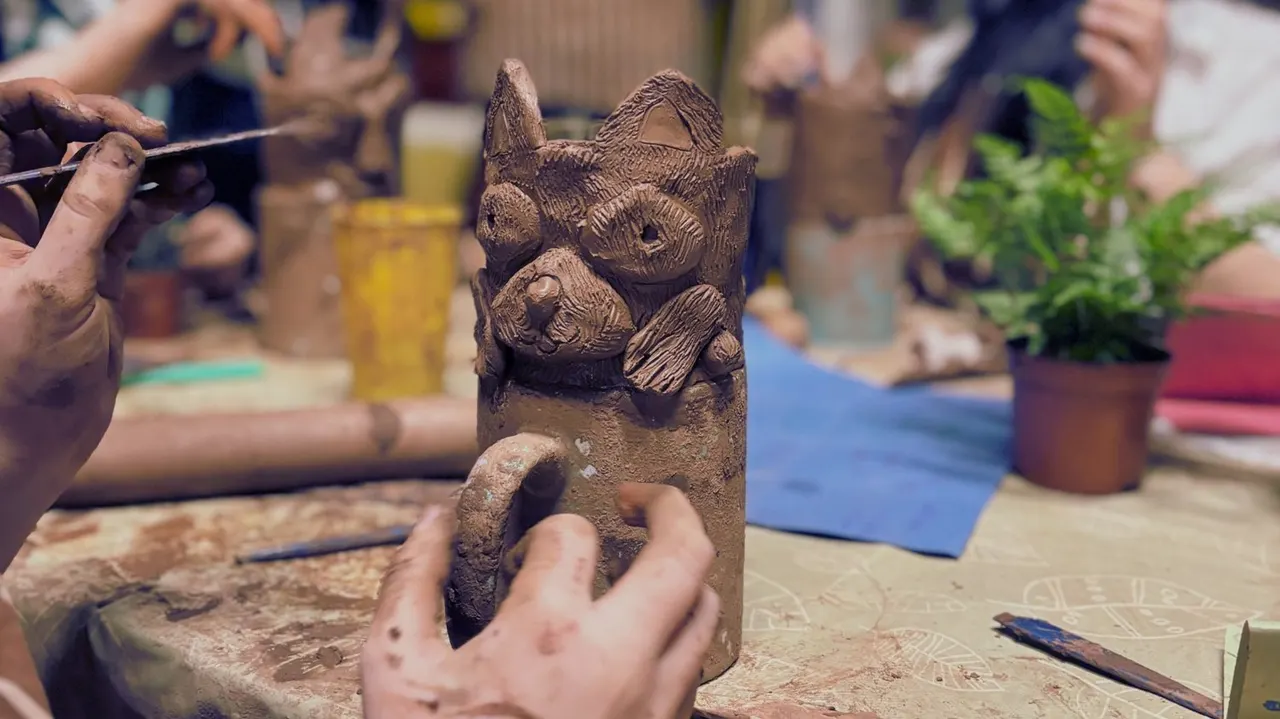 A person carving details into a clay sculpture of a cat with a paintbrush whilst others work on pottery nearby.