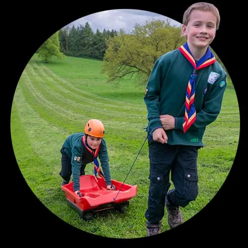 Two children in scout uniforms, one wearing an orange helmet seated in a red sledge being pulled by the other across a grassy hillside.