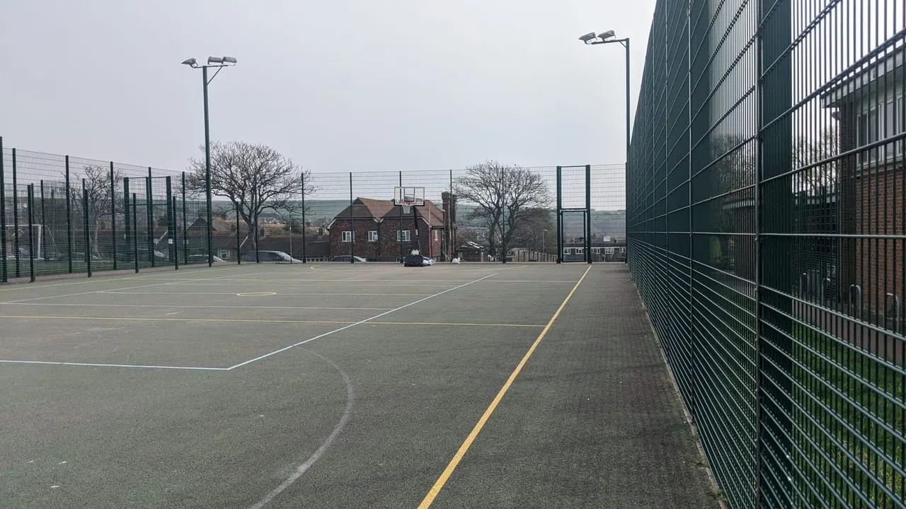 Empty outdoor sports courts with fenced enclosures, floodlights, and a brick building visible beyond the perimeter fence.