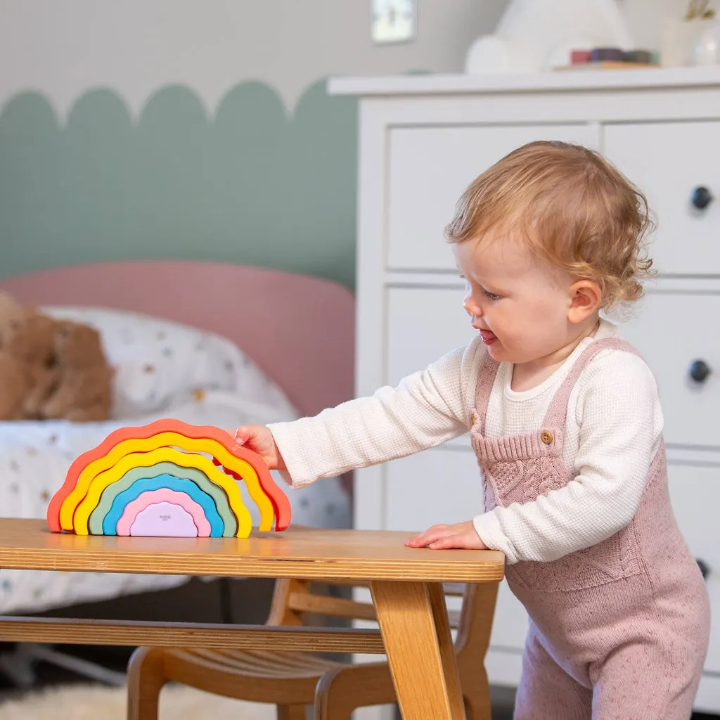 Toddler playing with a colourful wooden rainbow stacking toy at a table in a child-friendly room.