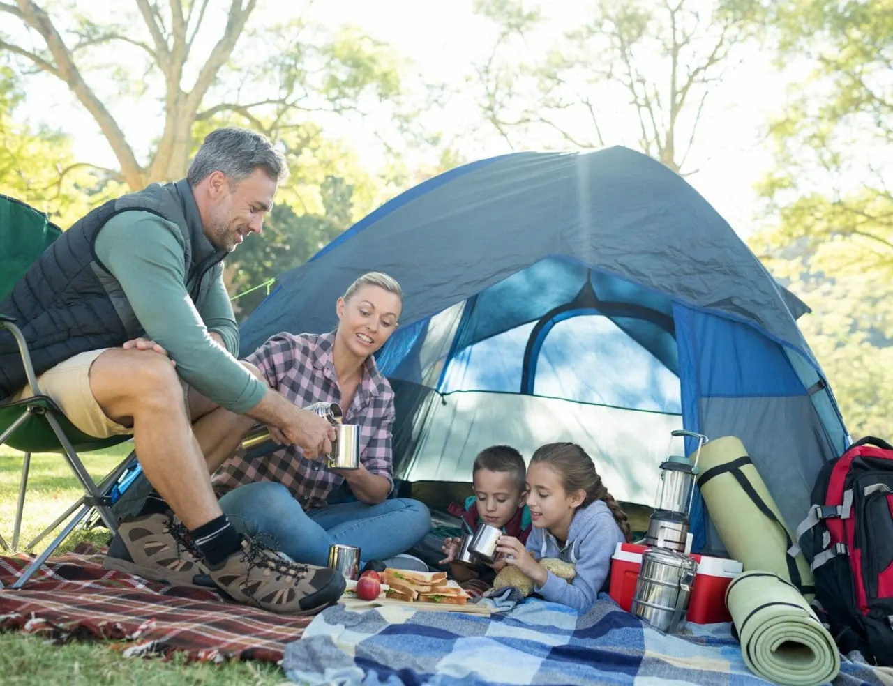 Family camping outdoors with two children eating inside and outside a blue tent, surrounded by trees.