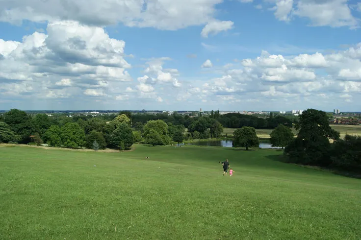 Visitors walking across open grassland with trees and a lake visible in the distance under a cloudy sky.