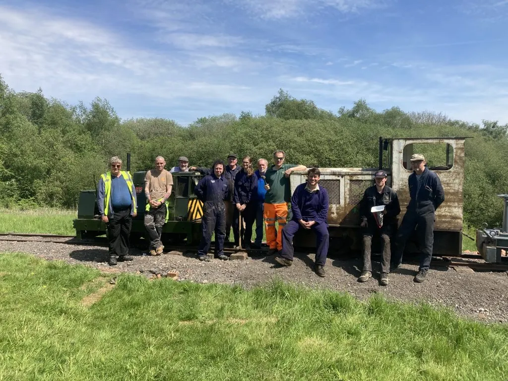 A group of visitors and staff pose together beside narrow-gauge railway locomotives at a heritage railway site, with woodland in the background.