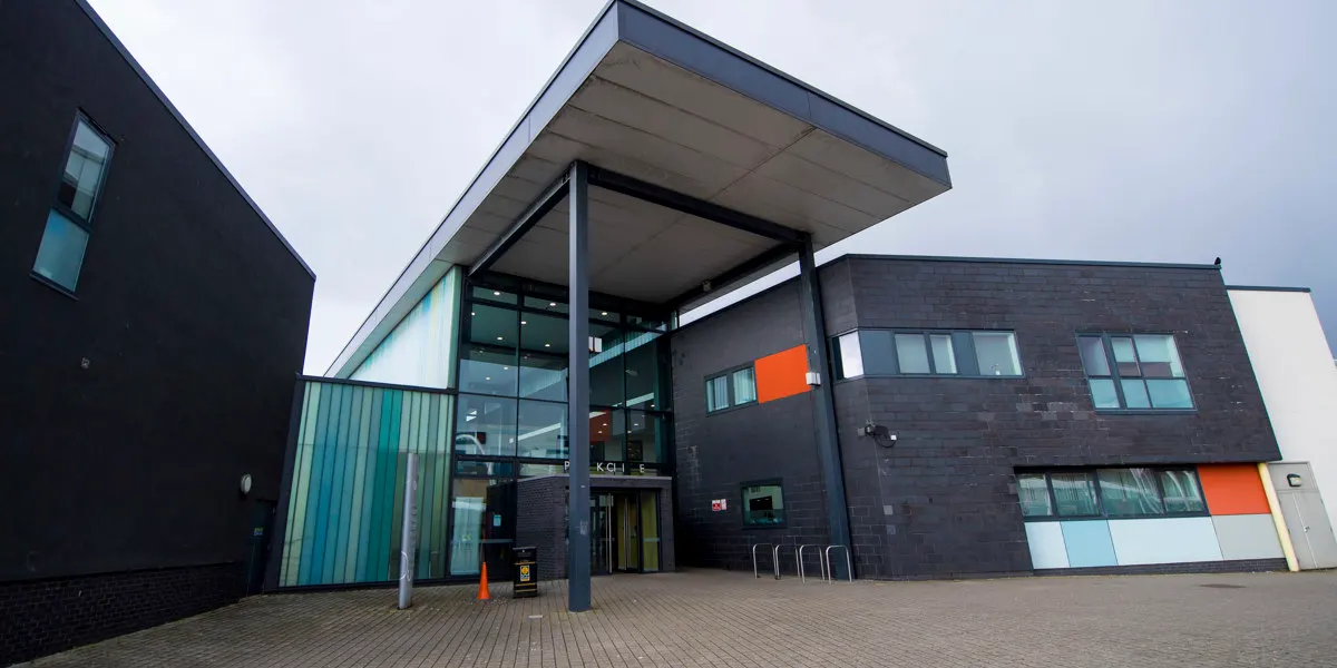Modern brick and glass leisure centre building with cantilevered roof and orange accents, exterior entrance courtyard.