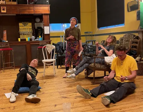 A group of five people participate in a drama workshop or rehearsal on a wooden stage floor.