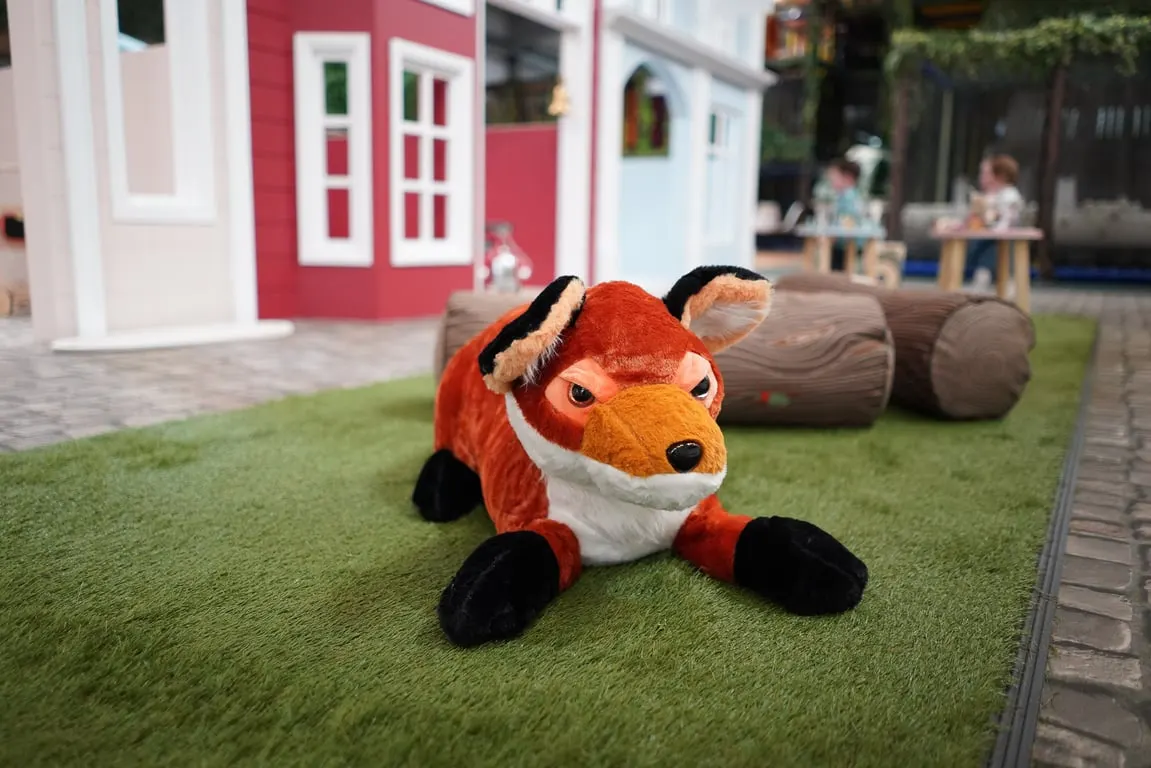 Soft toy fox on artificial grass with colourful play buildings and wooden logs behind it in an indoor play area.