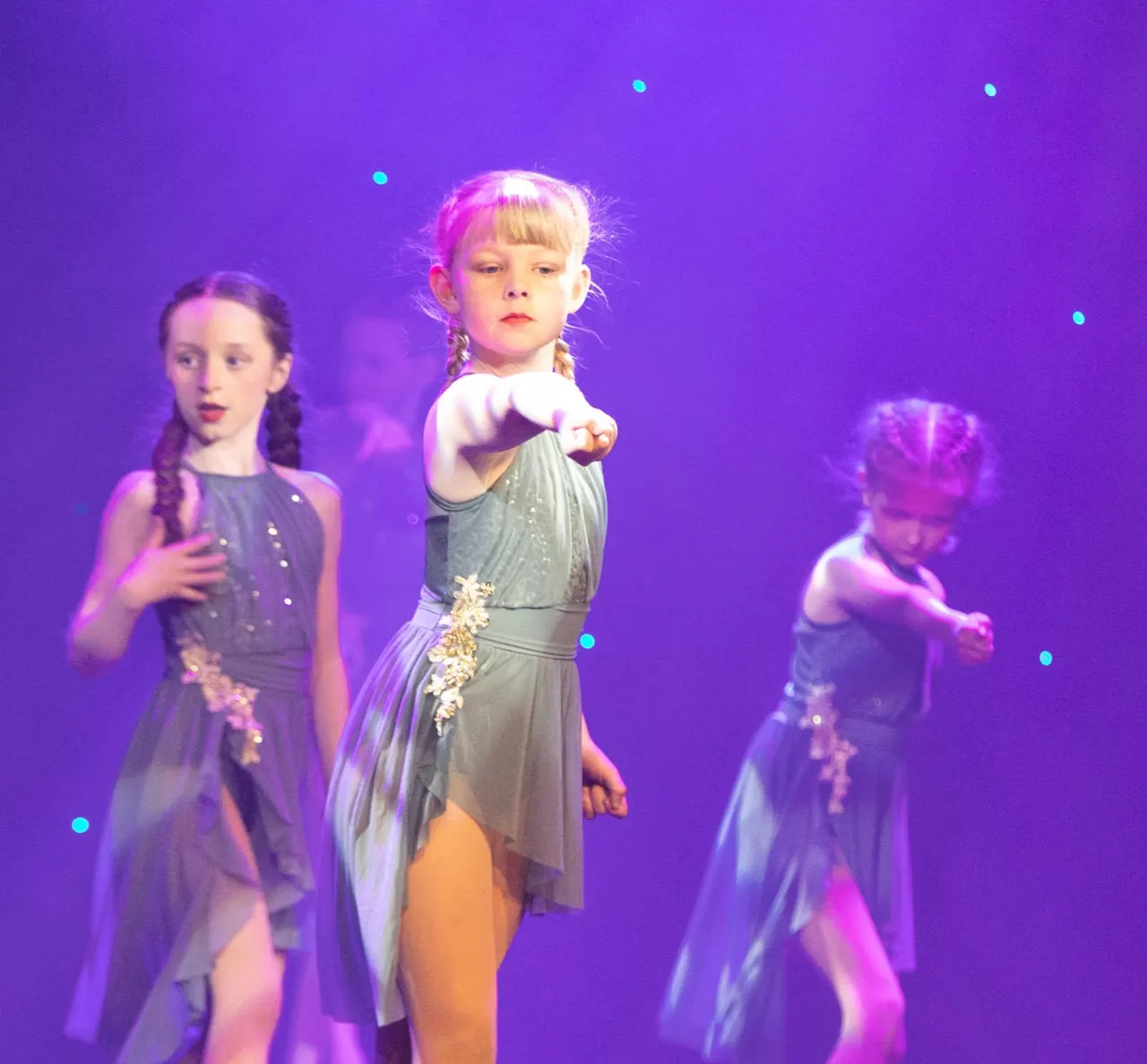 Three young children performing a dance routine on stage under purple lighting with sequinned costumes and cyan light effects.