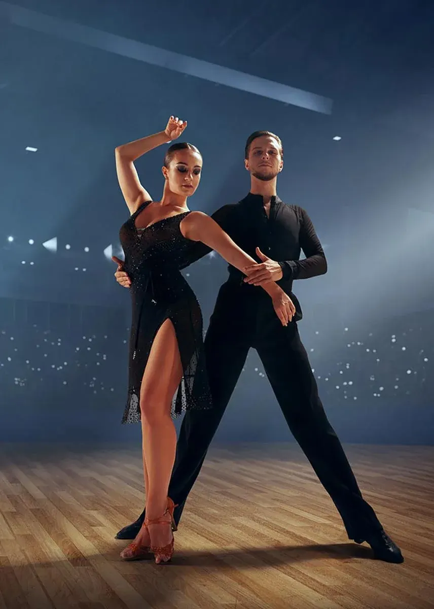 Two ballroom dancers in elegant black attire pose together on a wooden dance floor with atmospheric blue lighting.