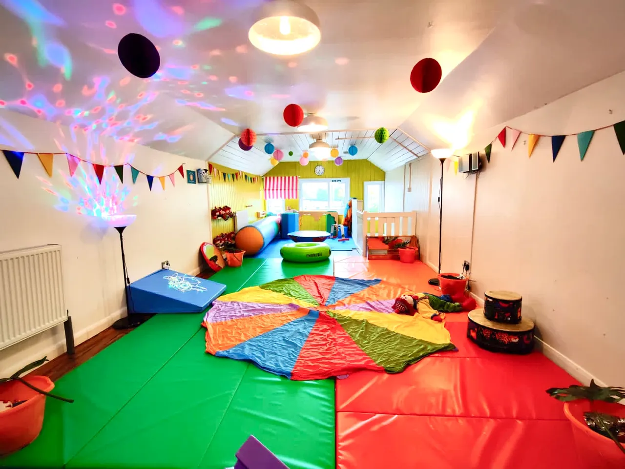 Colourful indoor play room with padded floor mats, parachute, balls, light projections and bunting decorations.