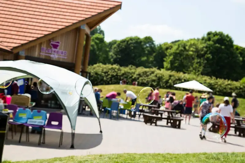 Outdoor leisure venue with wooden building, visitors seated at tables and picnic benches under shade, green fields and trees beyond.