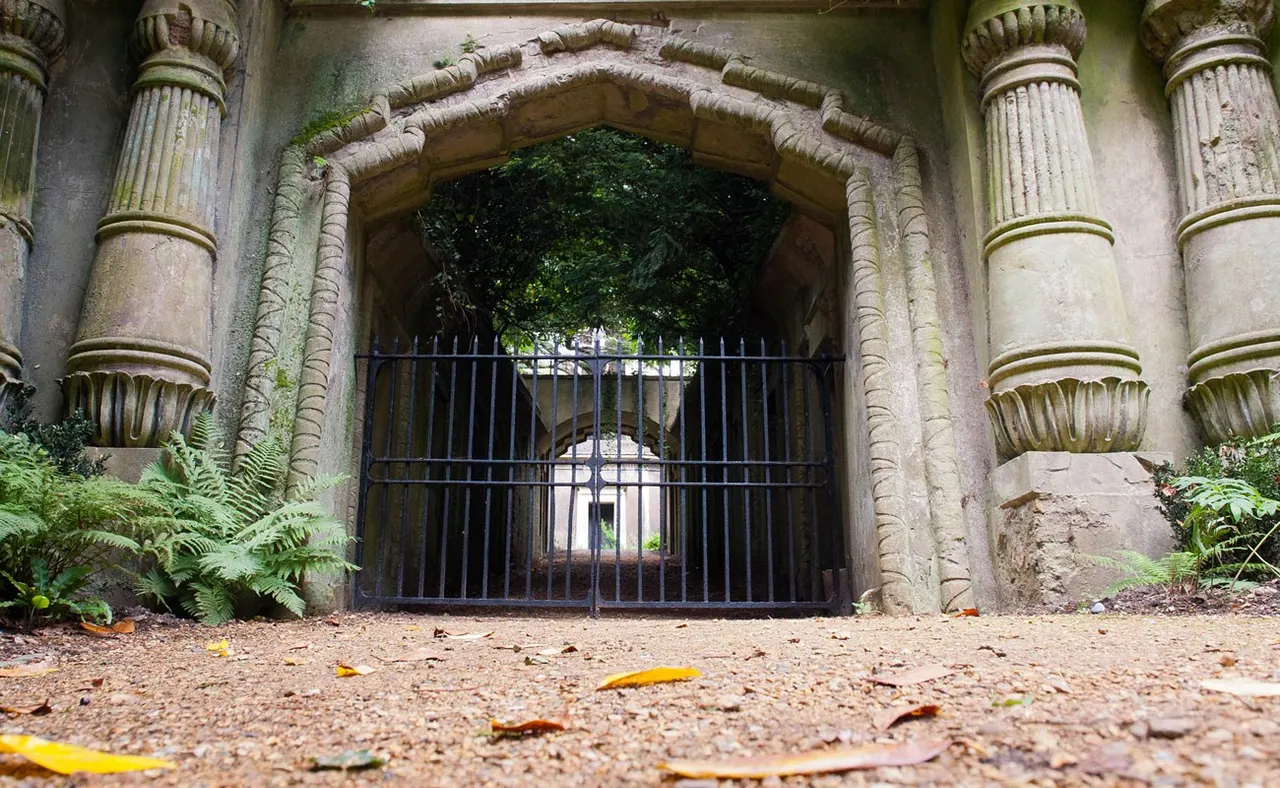 Ornate stone archway with decorative columns and iron gates opening onto a tree-lined path in a historic cemetery.