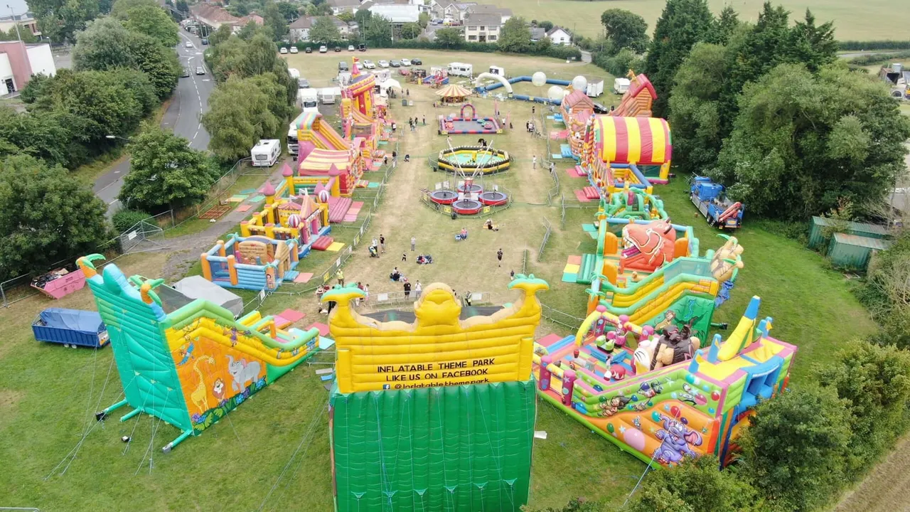 Aerial view of an inflatable theme park with colourful bouncy castles, slides and rides set up on a grassy field surrounded by trees and residential buildings.