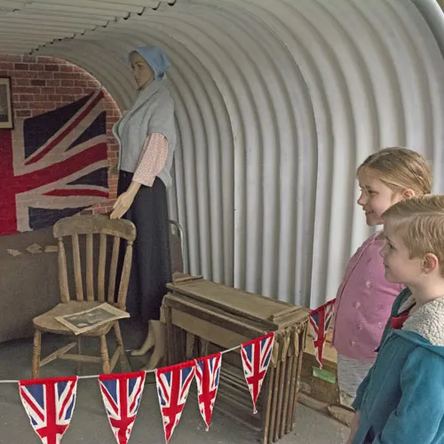 Two children viewing a wartime display featuring a figure and Union Jack bunting inside a corrugated metal shelter.