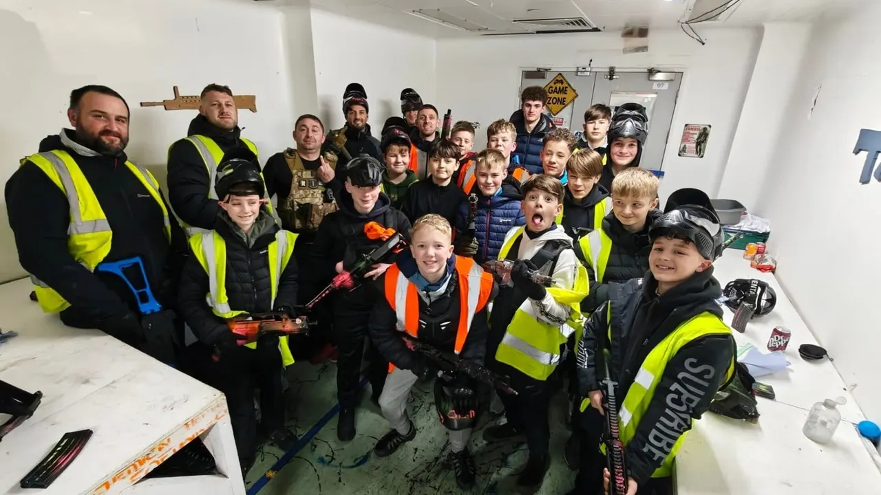 Group of children and adults wearing high-visibility vests and tactical gear posing together in an indoor airsoft game zone.