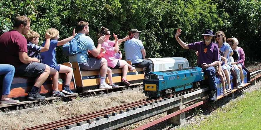 Adults and children riding in colourful open-air miniature train carriages along narrow-gauge tracks surrounded by greenery.