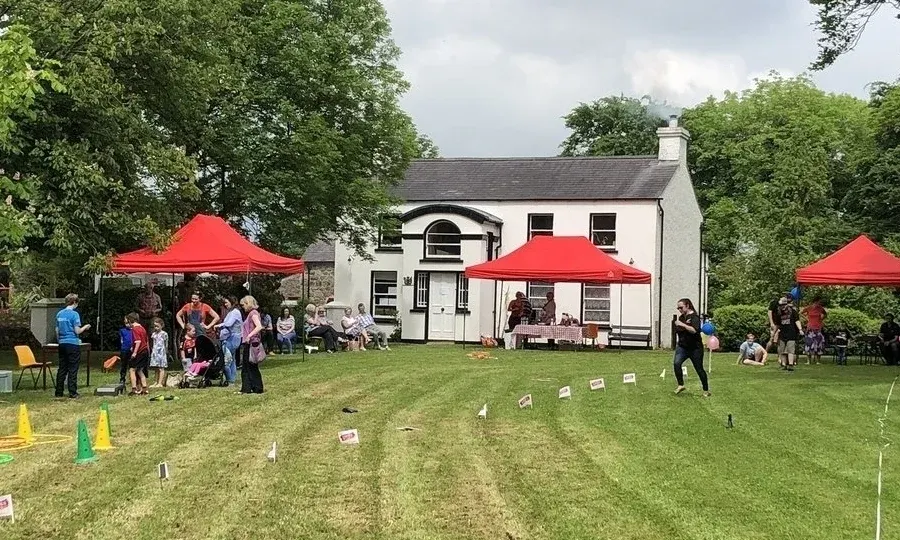 Children and adults at a garden party with outdoor games on the lawn, red marquees, and a white house with slate roof in the background.