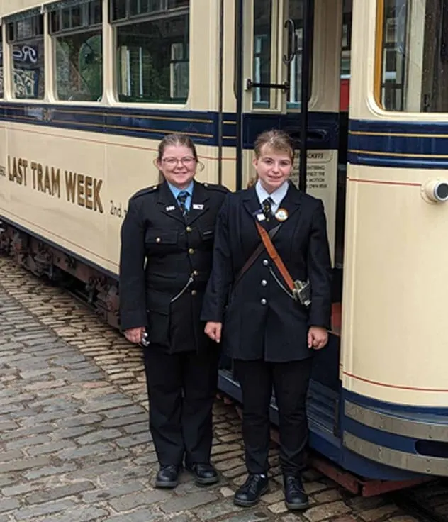 Two young people in uniform stand together next to a vintage tram.
