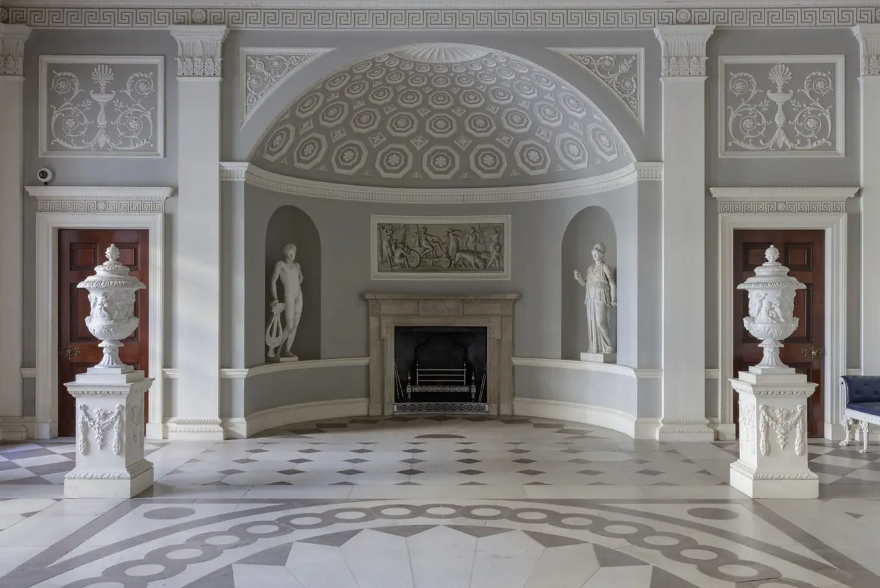 Neoclassical interior with domed ceiling, marble floor, fireplace, classical sculptures and ornamental urns.