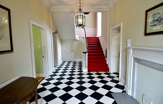 A grand hallway with black and white diamond-patterned floor, cream walls, white doors, and a red-carpeted staircase leading upwards.