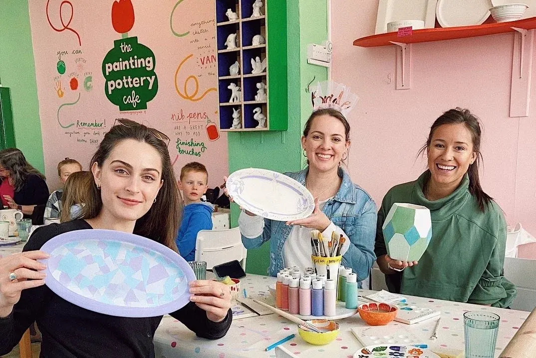 Three young people display hand-painted ceramic plates at a pottery painting activity table.
