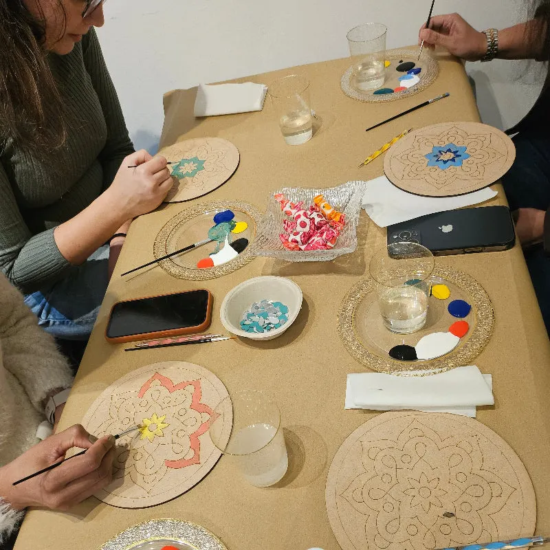 Two people paint designs on cork coasters at a table with paintbrushes, water glasses, and paint palettes.