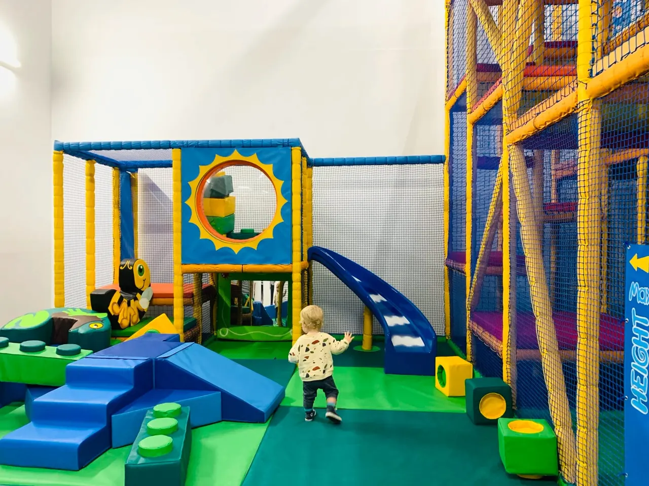 A toddler plays in a colourful soft play area with climbing structures, slides, and foam blocks.
