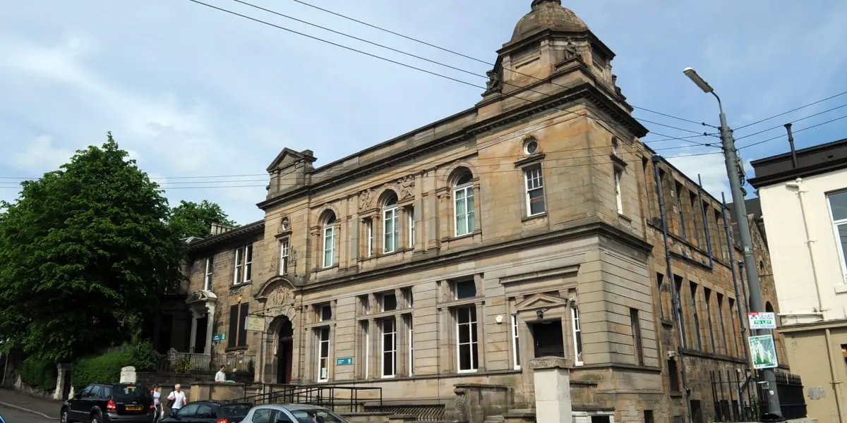Victorian stone library building with ornate architecture and central dome, parked cars and pedestrians on street outside.