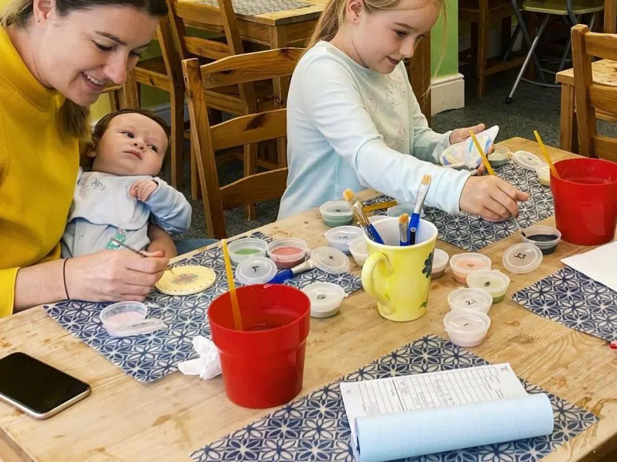 Two women and a toddler engaged in a pottery painting activity at a table with paints and brushes.