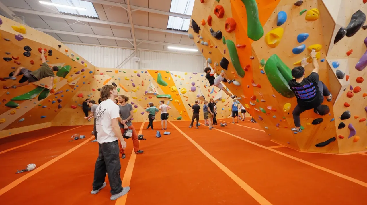 Multiple people rock climbing on colourful indoor climbing walls with an orange matted floor.