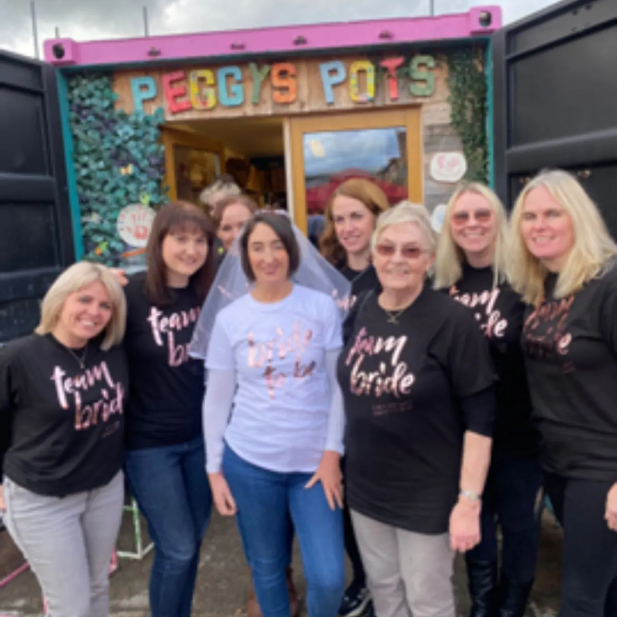 A group of women posing outside Peggy's Pots, most wearing matching black "team bride" t-shirts.