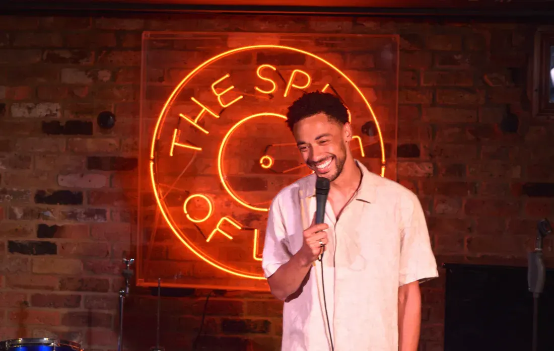 Man holding microphone performing stand-up comedy in front of illuminated neon sign on brick wall.