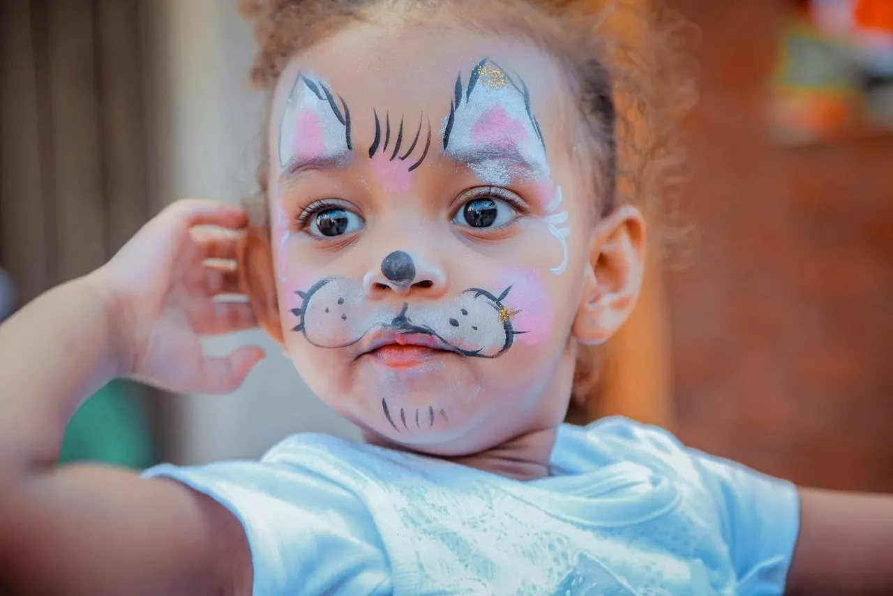 Young child with elaborate face paint designed as a cat with pink cheeks, blue eyes and whiskers.