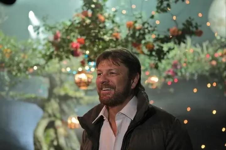 Man in black jacket smiling in front of festive floral and lights display indoors.