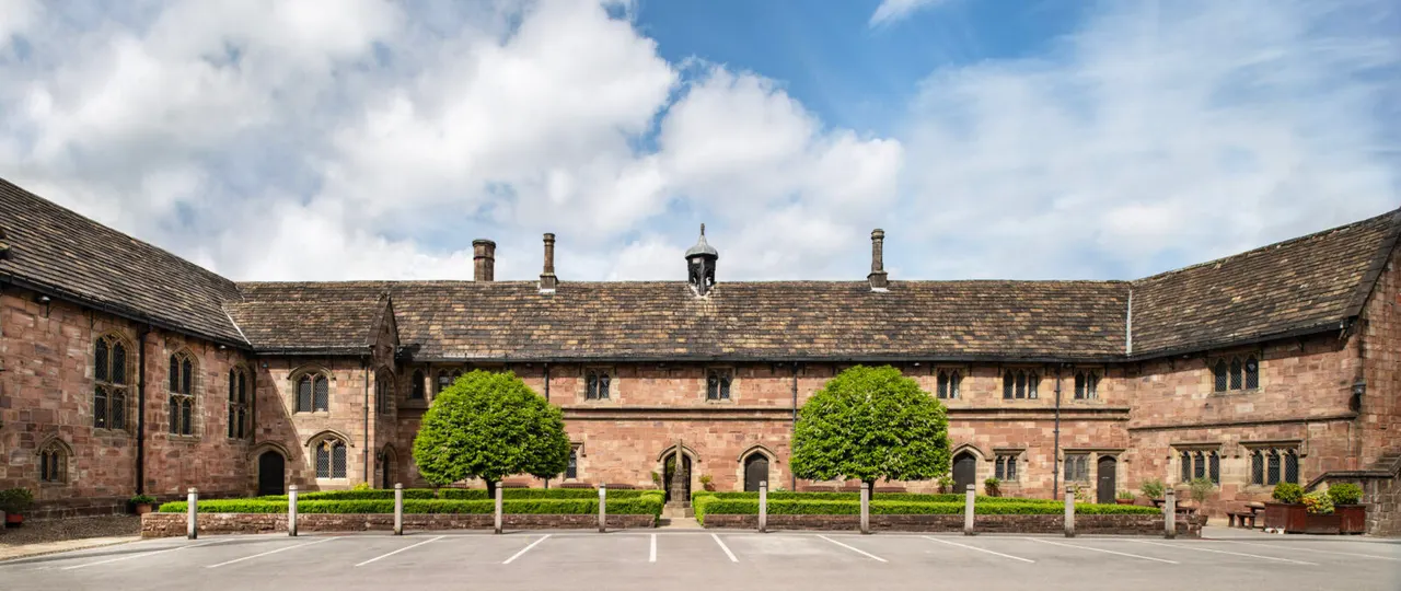 Historic stone building with red sandstone courtyard featuring manicured hedges and topiary trees under blue sky.