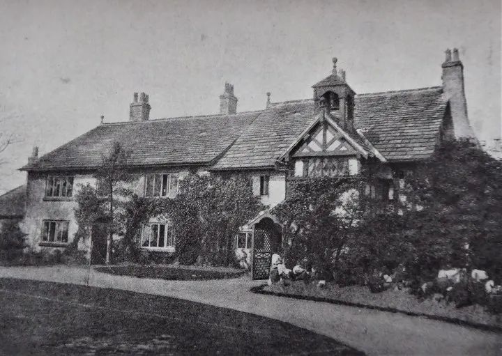 Historic black and white photograph of a half-timbered manor house with ivy-covered walls and multiple chimneys.
