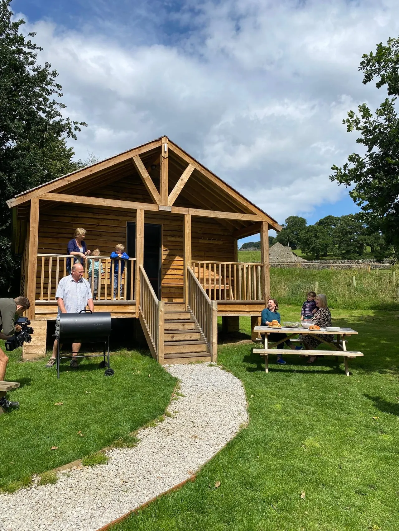 Families enjoying a wooden lodge with picnic tables and a barbecue in a grassy valley setting surrounded by trees.