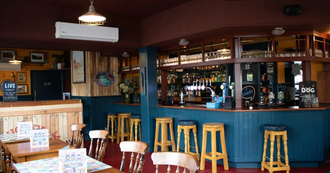 Empty pub interior with blue-painted bar, wooden bar stools, dining tables and chairs, shelves of bottles, and warm lighting.