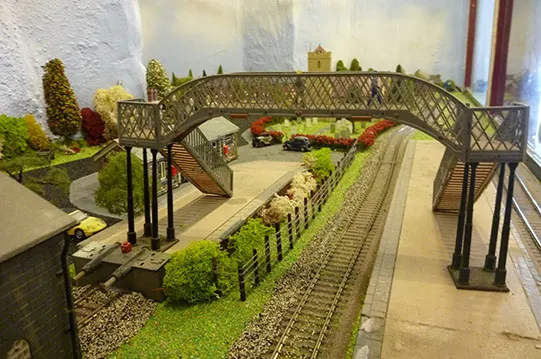 Detailed miniature railway layout with bridge, landscaping, and model buildings beneath archway.