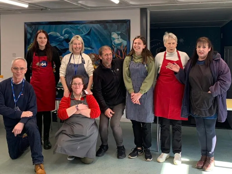 A group of seven adults and one person in a wheelchair pose together indoors, some wearing red aprons, in front of a blue mural.