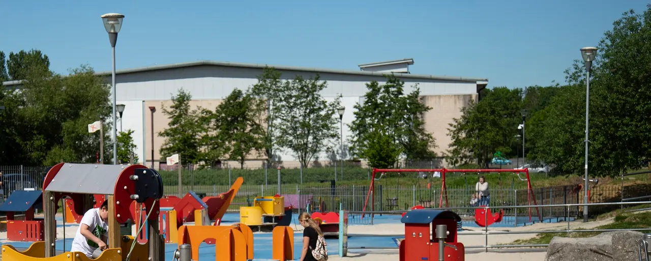 Children and adults enjoying a colourful playground with slides, swings and climbing equipment on a blue safety surface.