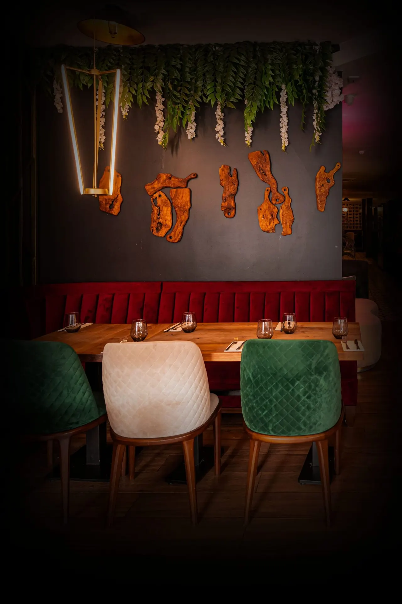 Restaurant dining booth with wooden table, colourful chairs, red upholstered banquette seating, and decorative orange wall art beneath hanging green foliage and neon light.