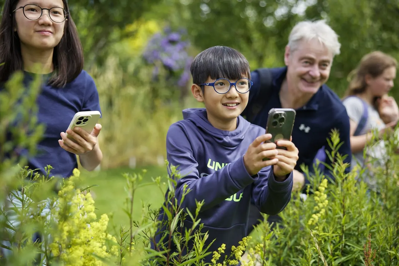 A young boy wearing glasses holds up a phone whilst standing amongst garden plants, with a teenage girl and older man beside him, all smiling in a lush green garden setting.