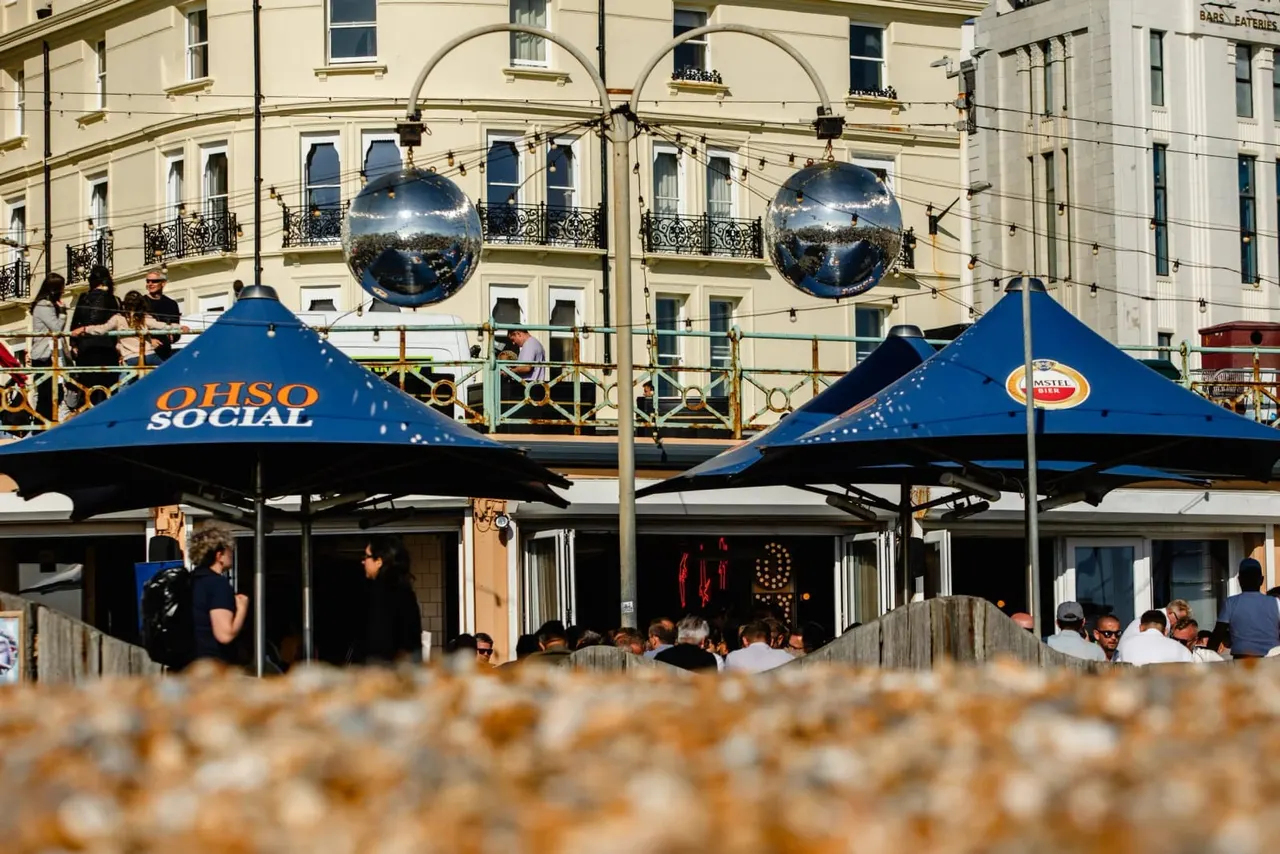 Beachfront bar and restaurant with blue canopy tents, diners seated outdoors, and ornamental spheres hanging above.