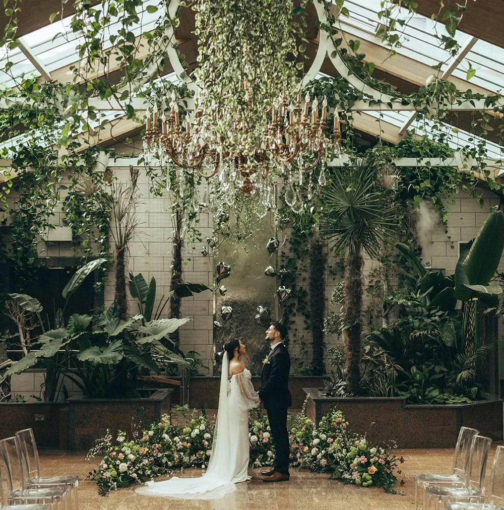 Bride and groom standing beneath a chandelier in a lush glasshouse surrounded by tropical plants and floral arrangements.